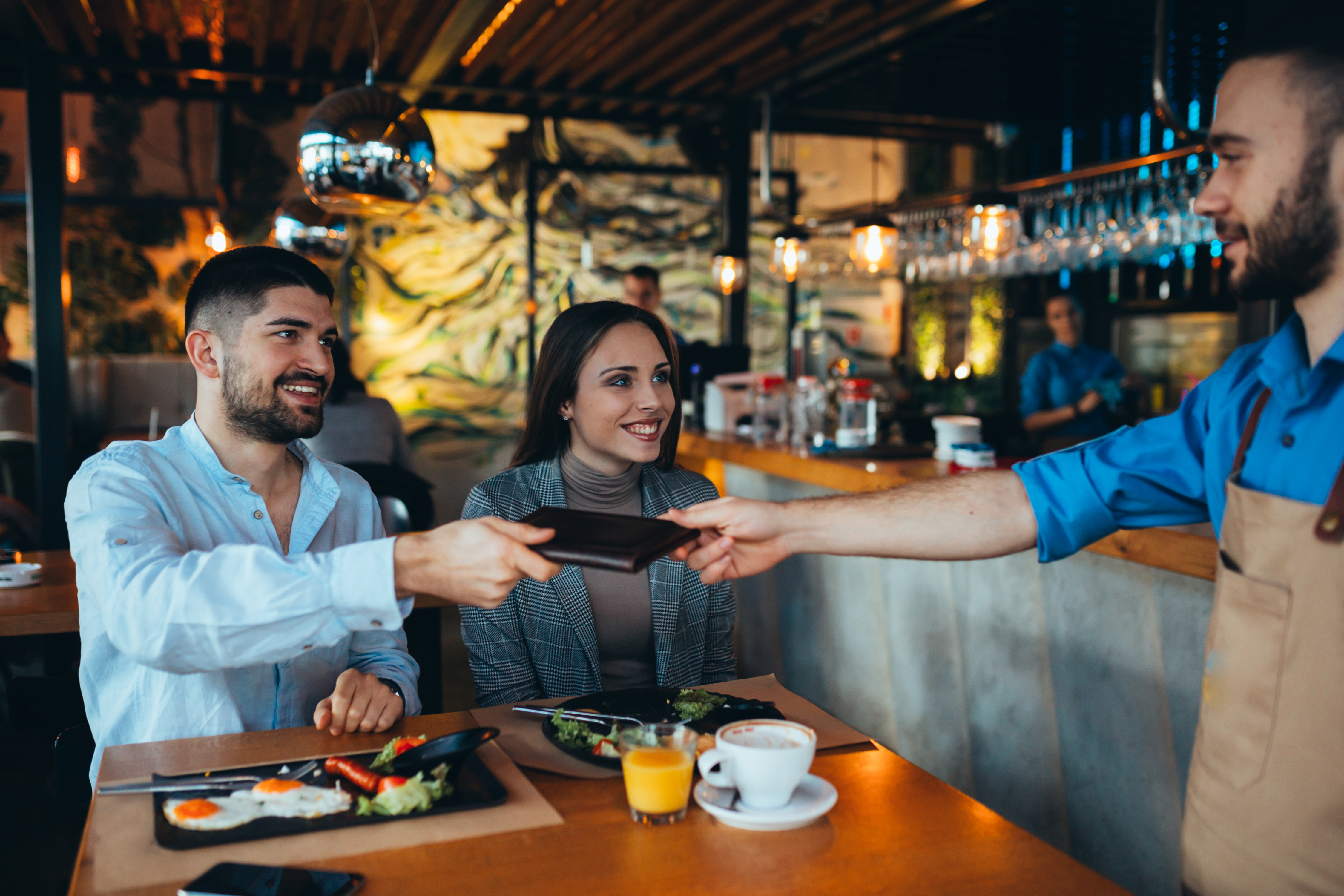 man smiling handing a waiter the check with a woman smiling next to him.